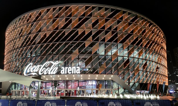 A large, modern arena with a distinctive rounded design featuring a complex pattern of illuminated panels. The facade displays the 'Coca-Cola arena' signage prominently. The venue is surrounded by barriers with 'World Tennis League' branding. The lighting creates a vibrant and inviting atmosphere at night, with lighting around the entrance and inside the building.